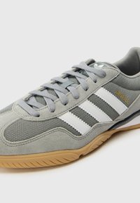 Grijze sport-schoen van mesh en suede materialen. Heeft drie witte strepen, grijze veters en een zool van gumrubber. Opvallende branding aan de zijkant.
