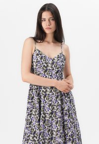 Blumenkleid mit tailliertem Oberteil, dünnen verstellbaren Trägern und einem ausgestellt Rock. Verziert mit lila, weißen und grünen Blumenmustern auf einem dunklen Hintergrund.