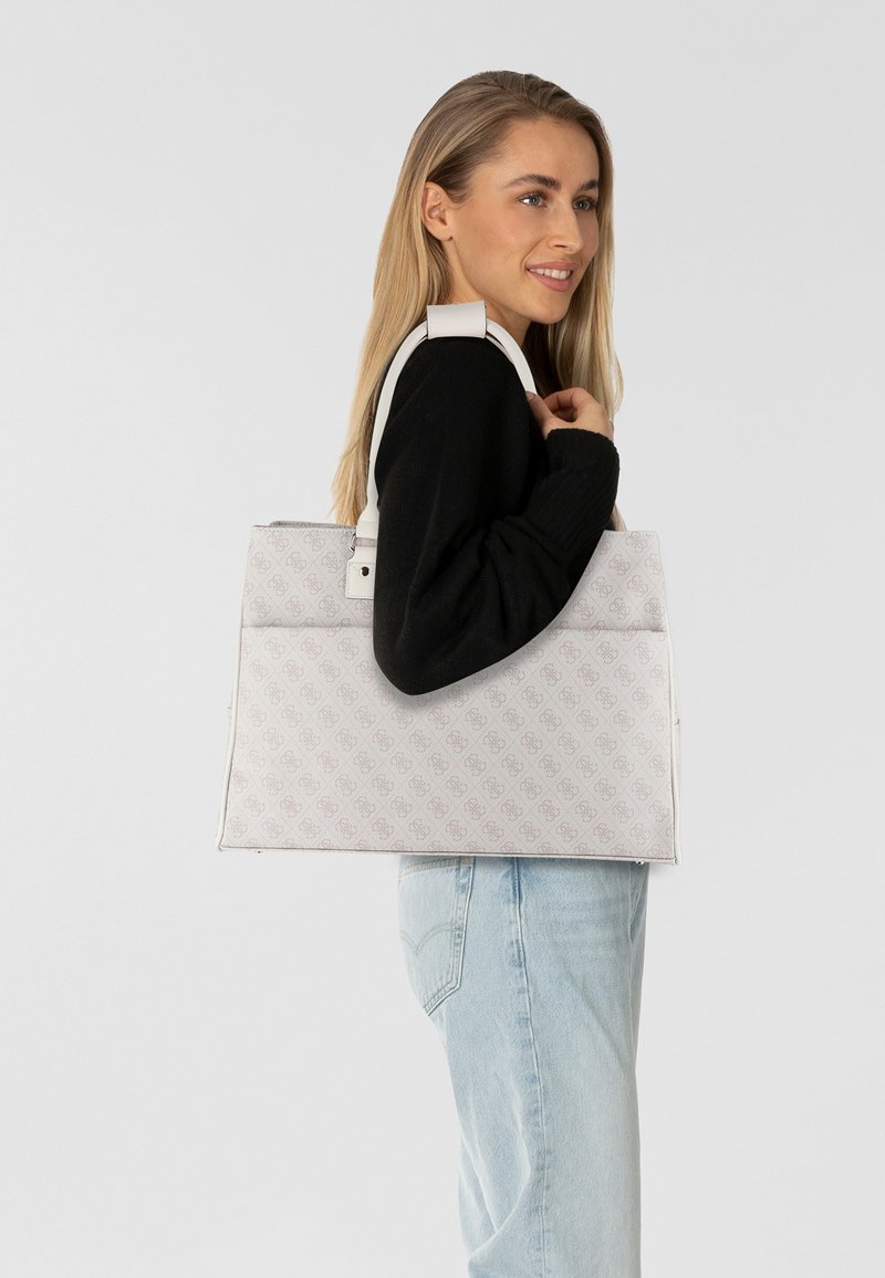Bolso tote blanco con un patrón de logotipo en relieve, forma estructurada, asas resistentes, textura suave y un atuendo negro en contraste que lleva puesto el modelo.