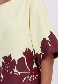 Crème-kleurig T-shirt met korte mouwen en grote donkerrode bloemen uitsnijdingen langs de zoom en de mouwranden.