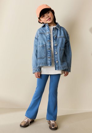 Mladá dívka s růžovou čepicí, nadměrně velkou džínovou bundou, bílým tričkem, modrými kalhotami a metalickými teniskami stojí u béžové zdi.