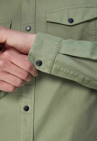 Camicia verde oliva in tessuto testurizzato, con polsini con bottone e bottoni a pressione neri. Due tasche sul petto aggiungono un tocco funzionale.