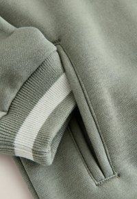 Zbliżenie na materiał w kolorze szałwii z mankietem z białym paskiem oraz boczną kieszenią na bluźnie lub kurtce.