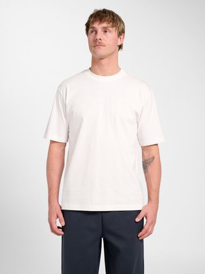 Man met kort haar en snor die een eenvoudig wit T-shirt met korte mouwen en een donkere broek draagt, staand tegen een lichtgrijze achtergrond.