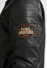Chaqueta de cuero negra con textura acolchada en los hombros y un parche bordado marrón con la marca "Petrol Industries".