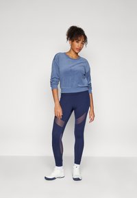 Frau mit lockigem Haar, die ein blaues Langarmoberteil, marinefarbene Leggings mit Netzpaneelen, weiße Socken und weiße Turnschuhe trägt und vor einem schlichten Hintergrund steht.