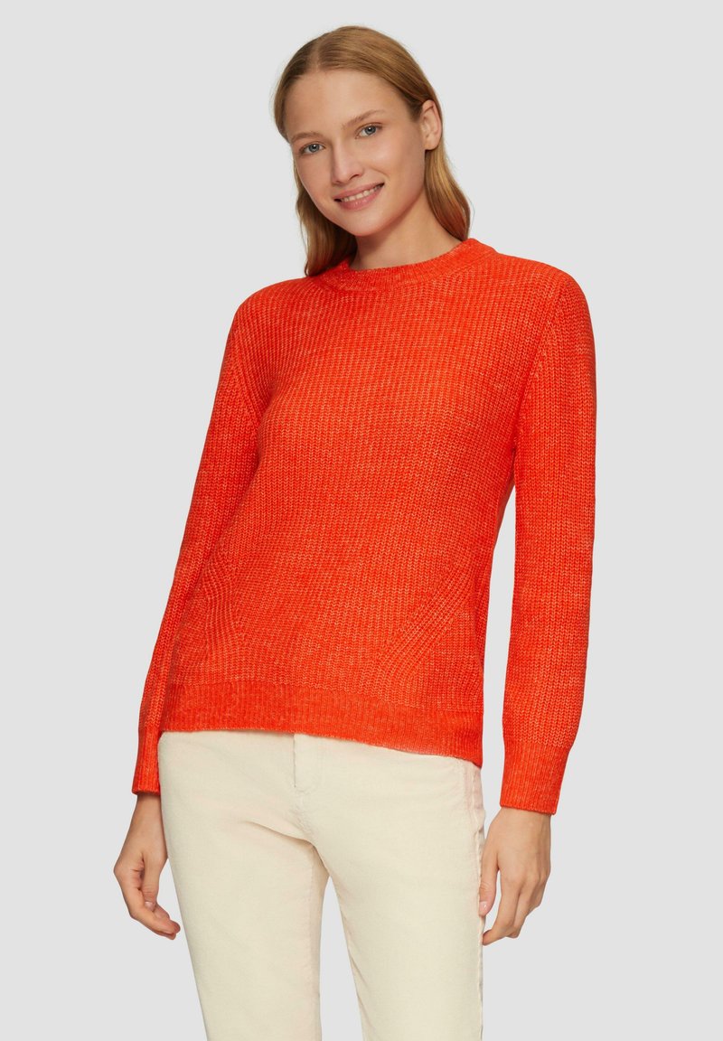 Helles orange gestricktes Sweatshirt mit rundem Halsausschnitt, langen Ärmeln und strukturierter Oberfläche. Über hellen Hosen getragen.