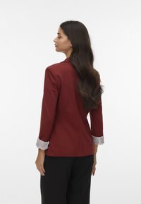 Czerwony blezer o dopasowanym kroju, z podwiniętymi paskami w poziome paski i gładką teksturą. Widok z tyłu ukazuje dopasowany kształt i długość.