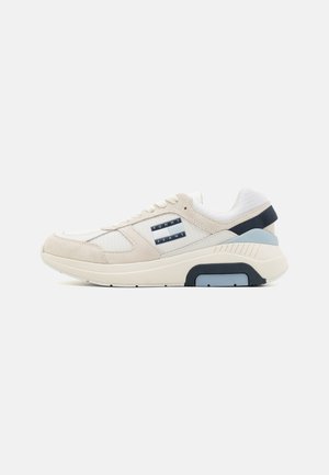 Zapatilla beige con una mezcla de materiales de ante y textil. Presenta una suela acolchada, cordones blancos y un acento en azul marino en el talón. Logo de la marca visible.