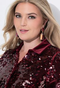 Blusa burdeos de lentejuelas con cuello de satén. Presenta grandes pendientes geométricos con acentos en rojo y naranja. Superficie lisa y detalles brillantes.