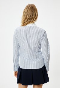 Camicia a righe blu chiaro e bianca con colletto classico, abbinata a una gonna plissettata blu navy. Presenta maniche lunghe e dettagli con bottoni.