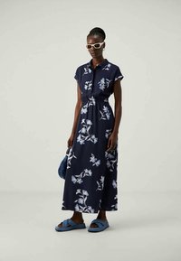 Vestido maxi florido azul-marinho com mangas curtas, cintura ajustada com cinto, padrões de flores em azul claro, combinado com sandálias deslizantes azuis e uma bolsa azul.