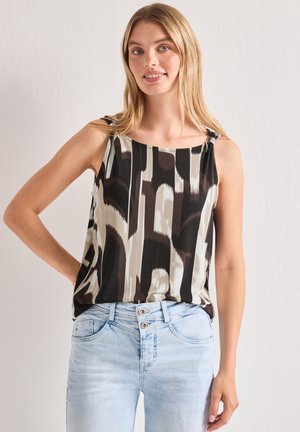 Lyshåret kvinde iført en ærmeløs sort, brun og beige mønstret top og lyseblå jeans med dobbelt knap, stående op ad en ensfarvet væg.