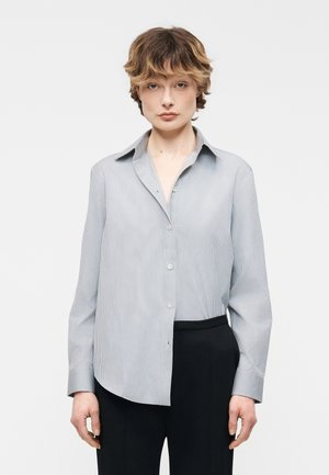 Osoba z krótkimi, falowanymi włosami, ubrana w jasnoniebieską koszulę w cienkie paski na guziki oraz czarne spodnie, stojąca na tle jednolitej białej ściany.