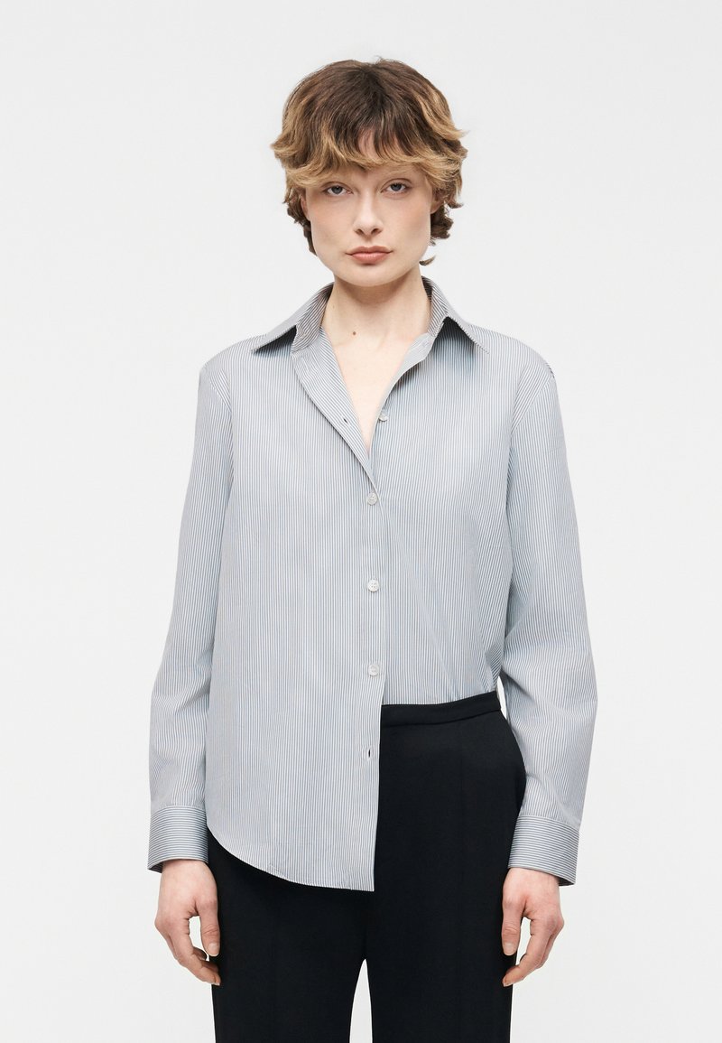 Άτομο με κοντά κυματιστά μαλλιά, φορώντας ένα ανοιχτό μπλε πουκάμισο με ρίγες και μαύρο παντελόνι, στέκεται μπροστά σε απλό λευκό φόντο.