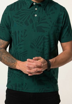 Homme portant un polo vert foncé avec un motif de feuilles noires, bras gauche tatoué, bracelet noir, mains jointes devant.