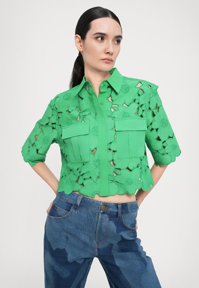 Green cropped shirt featuring floral cut-outs, two front pockets, and a collared neck. Worn with blue denim jeans.