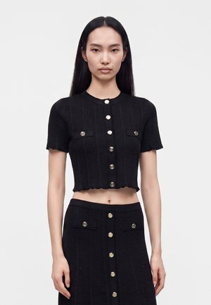 Ung kvinde med langt sort hår bærer en sort kortærmet knaplukket crop top og matchende højtaljet nederdel med guldknapper.