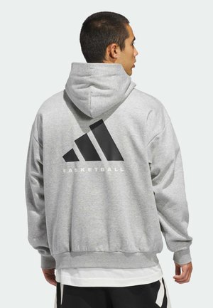 Mann in grauem Kapuzenpullover mit großem schwarzem Adidas-Logo und dem Wort "BASKETBALL" auf der Rückseite, seitlich stehend.