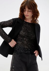 Μαύρη μπλούζα με παγιέτες και μακριά μανίκια, φορεμένη κάτω από μαύρη σαλοπέτα. Το σύνολο συνδυάζεται με σκούρα, φαρδιά τζιν παντελόνια που διαθέτουν λεπτομέρειες κορδονιού.