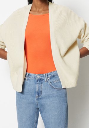 Femme portant un haut orange, un cardigan beige clair ouvert devant, et un jean bleu clair taille haute, les mains sur les hanches.