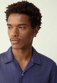 Navy blue short-sleeve shirt with a textured pattern, featuring a collar and a single button at the neckline. Smooth material and relaxed fit.