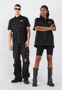 Camicia nera a maniche corte con cuciture bianche e due tasche sul petto. Abbinala a jeans neri strappati e stivali neri fino al ginocchio con dettagli metallici.