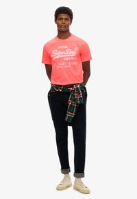 T-shirt rose néon avec texte blanc, jean noir avec les ourlets retroussés, et baskets beige. Chemise à carreaux verte et rouge nouée à la taille.