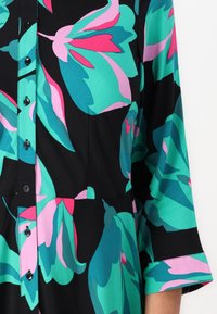 Värgglad blommig button-up klänning med stora teal-, rosa- och gröna mönster på en svart bakgrund; mjukt tyg med trekvartslånga ärmar.