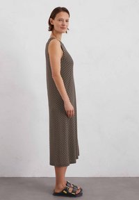 Ärmelloses Maxikleid in Braun mit geometrischem Muster, aus weichem Stoff gefertigt. Es hat eine lockere Passform und einen geschwungenen Saum, kombiniert mit schwarzen Sandalen.