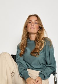 Tirkīza krāsas, teksturēts džemperis ar brīvi krītošām piedurknēm un ribotām aprocēm, kombinēts ar bežām, augstās jostasvietas biksēm.