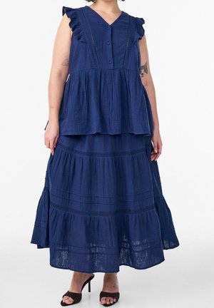 Femme portant une robe bleu marine sans manches à étages avec des détails à volants et des talons hauts ouverts noirs, debout sur un fond blanc.