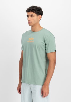 Mann trägt ein hellgrünes Alpha Industries T-Shirt mit orangefarbenem Logo, kombiniert mit hellblauen Jeans, stehend vor weißem Hintergrund.