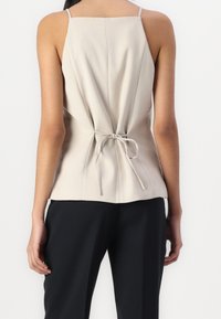 Top sin mangas beige con la parte posterior ajustada atada con una cinta delgada, que presenta una textura suave y un diseño estructurado. Combinado con pantalones negros.