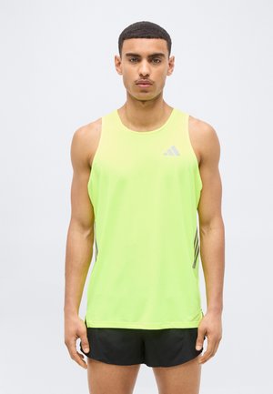 Noor mees, kes kannab neoonkollast varrukateta sporditoppi ja musti pükse, seisab näoga otse vaataja poole ühetaustalise tausta ees.