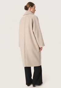 Palton bej dintr-un amestec de lână, cu un guler larg, umeri lăsați și o croială lejeră, asortat cu pantaloni negri evazați. Textură netedă și design minim.
