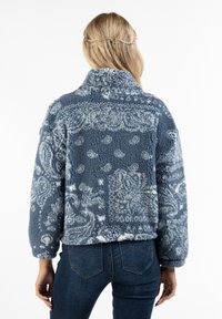 Fleecejakke i blå med et tekstureret blomstermønster, høj krave og lange ærmer; har en cropped silhuet og blød tekstur.
