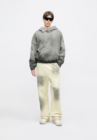 Γκρίζα oversized φόρμα με ξεθωριασμένη υφή, συνδυασμένη με εκρού παντελόνι που έχει βαθμιαία σκοτεινές κηλίδες. Το μοντέλο φοράει σκούρα γυαλιά ηλίου.