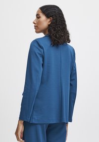 Blaues Langarmoberteil aus einem weichen Stoff, mit geradem Schnitt und seitlichen Schlitzern, mit einem dezenten Kragen und sauberen Nähten.