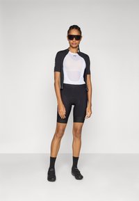 Cycling outfit featuring a black short-sleeve jersey and padded black shorts. The jersey has a white mesh panel, and the model wears sunglasses.