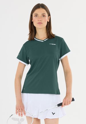 Kvinde iført en mørkegrøn sportstrøje og hvid nederdel, der holder et badmintonketsjer i højre hånd mod en ensfarvet baggrund.