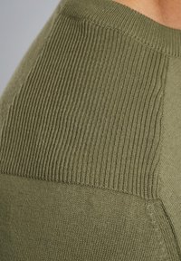 Olivno zelena jopica z rebrasto teksturo na ramenu in gladkim delom telesa, ki poudarja kontrastne vzorce tkanin.