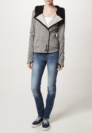 Woman wearing a grey zip-up hoodie with black lining, blue slim-fit jeans, and navy sneakers with white laces, standing against a plain background.