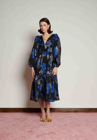 Γυναίκα που φοράει μαύρο μίντι φόρεμα με μπλε και χρυσά λουλουδάτα σχέδια, φουσκωτά μανίκια, χρυσά κοσμήματα και χρυσά ψηλοτάκουνα παπούτσια με μυτερή μύτη, στέκεται σε ροζ χαλί.