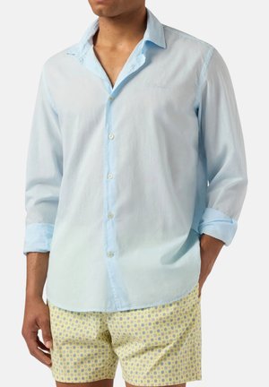 Uomo che indossa una camicia celeste a maniche lunghe con bottoni e pantaloncini gialli con motivo, con una mano in tasca su sfondo uniforme.