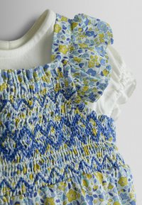Hvid babytrøje under blå og gul blomsterkjole med smocket krop og flæseærmer på ensfarvet baggrund.