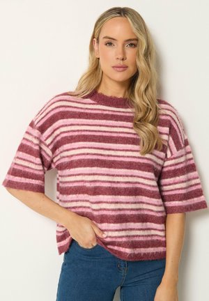 Kvinde med langt blond hår iført en lyserød og mørkerød stribet kortærmet sweater og blå jeans, stående med den ene hånd i lommen.