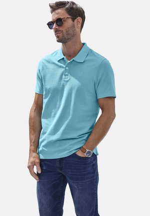 Türkises Polohemd mit Kragen und zwei Knöpfen, kombiniert mit dunkelblauen Jeans. Das Model trägt schwarze Sonnenbrillen und eine braune Uhr.