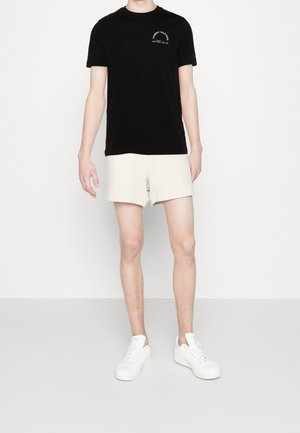 Musta pyöreäpääntieinen puuvillainen T-paita, jossa valkoinen logopainatus, yhdistetty beigenvärisiin pellavashortseihin. Malli käyttää valkoisia tennareita.