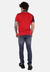 Polo rosso con maniche corte, dettagli blu navy e colletto bianco. Indossato con jeans blu e scarpe nere, in uno stile casual.